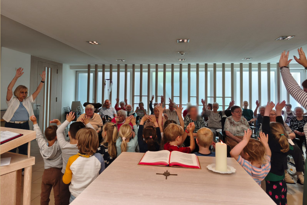 Eine bunte Gruppe aus Kindern und älteren Menschen versammelt sich in einem hellen, modernen Raum mit großen Fenstern und Holzvertäfelung. Alle Teilnehmer heben die Hände. Im Vordergrund befindet sich ein mit einem offenen Buch, einer weißen Kerze auf einem Teller, einem holzfarbenen Kreuz.