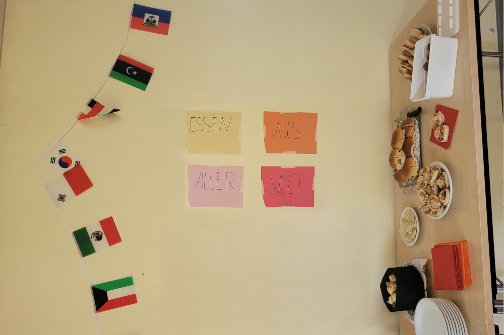 Buffet mit internationalen Speisen. Darüber hängen Flaggen verschiedener Länder und ein Schild „Essen aus aller Welt“.