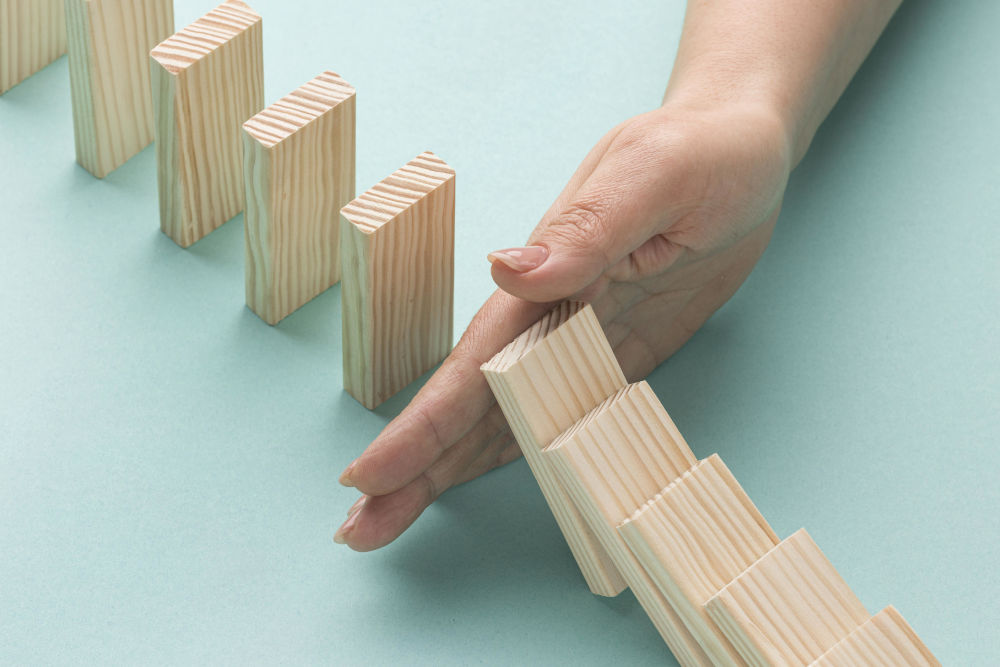 Eine Hand stabilisiert einen Holzklotz in einer Reihe von aufsteigenden Klötzen, die wie eine Stütze oder ein Domino-Effekt angeordnet sind. Das Bild symbolisiert präventives Handeln